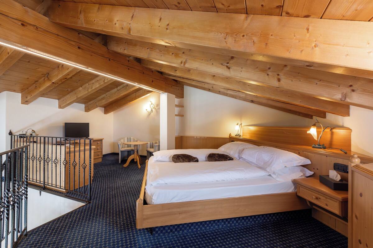 A wooden ceiling with exposed beams, a bed with white linens, a bedside table with a lamp, and a small desk with a chair.