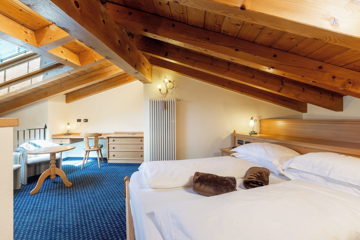 A bedroom with a sloped ceiling, a bed with white linens, a desk with a chair, and a radiator.