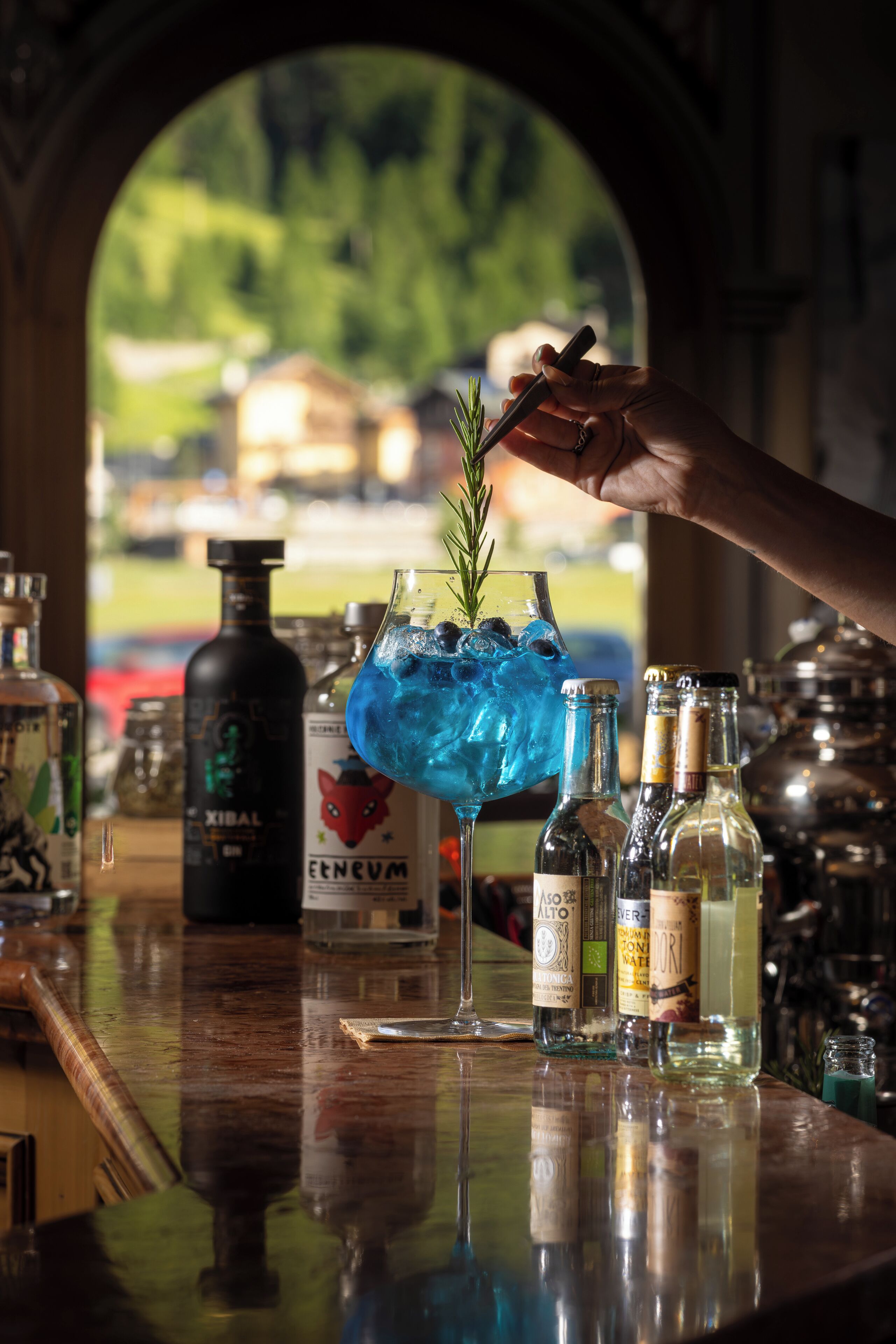 Un bar con una variedad de botellas y un cóctel azul adornado con una ramita de romero.