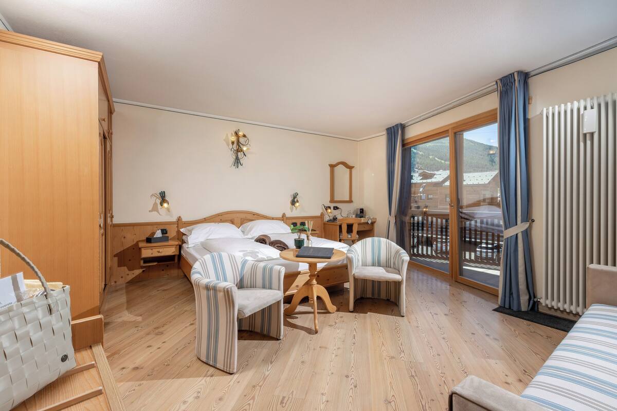 A hotel room with a bed, two chairs, a small table, a wooden wardrobe, a nightstand, a mirror, and a view of the outdoors through a large window.