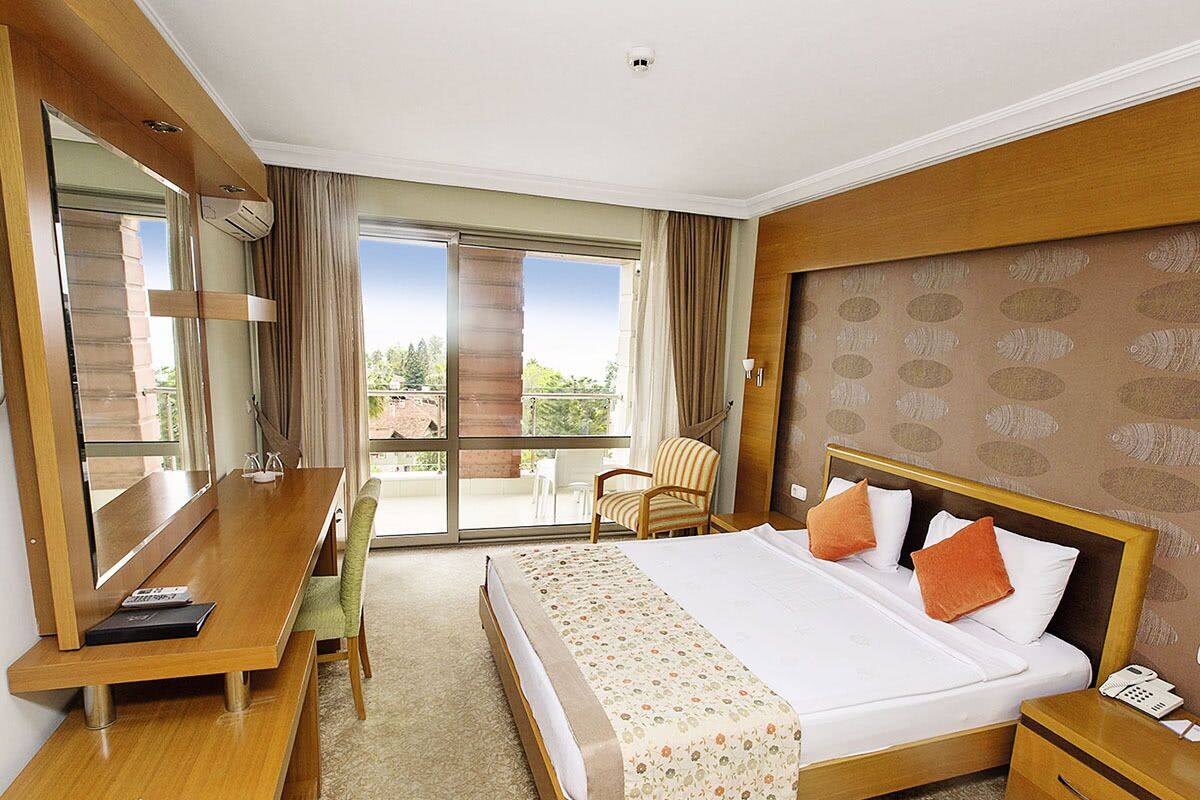 A hotel room with a bed, a desk, a chair, a bedside table, and a large window with a view.