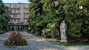 A hotel entrance with a central flower bed, flanked by statues, and surrounded by lush greenery. - Hotel Terme Villa Piave (Abano Terme)