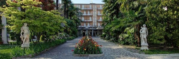 A hotel entrance with a central flower bed, flanked by statues, and surrounded by lush greenery.