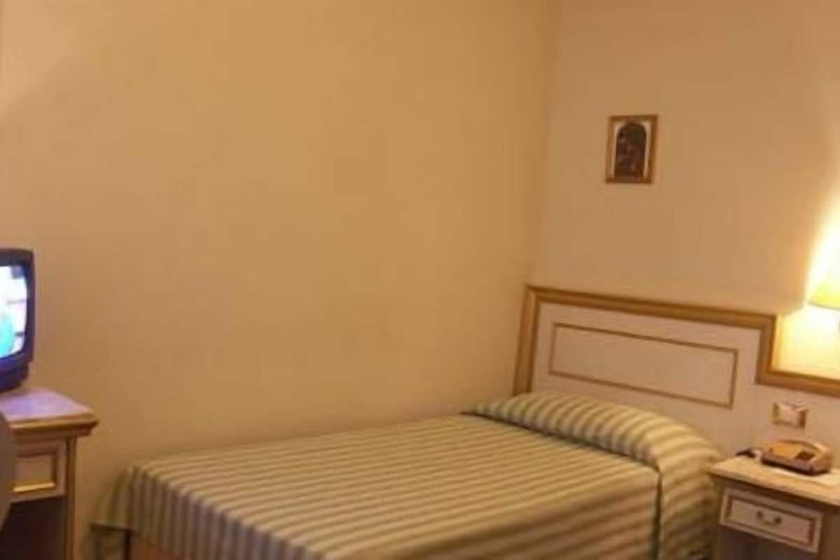 Une chambre d’hôtel avec un lit simple, un téléviseur posé sur une table de chevet, une lampe et un petit tableau accroché au mur.