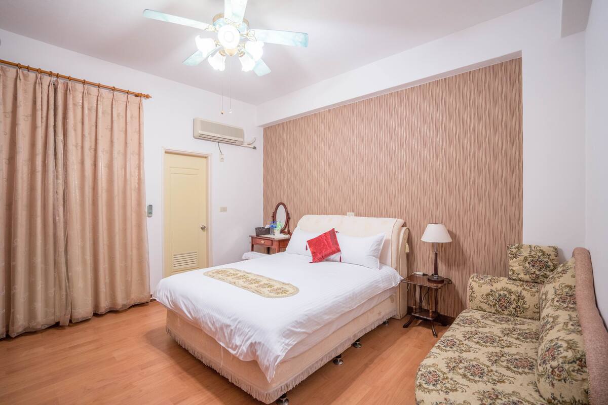 A bedroom with a bed, a nightstand, a lamp, a sofa, and a ceiling fan.