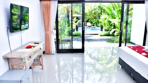 A modern hotel room with a large sliding glass door leading to a pool area, a flat-screen TV mounted on the wall, a wooden dresser, and a bed with a red and white bedspread. - Palm Garden Bali (Nusa Dua)
