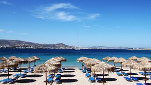 A beach with thatched umbrellas and blue lounge chairs, a sailboat in the distance, and a clear view of the coastline. - Paradiso Boutique Hotel (Paros)