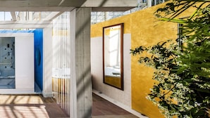 Een smalle gang met een glazen deur die naar een kamer leidt, een gele wand met een lijst en een plant aan de rechterkant.