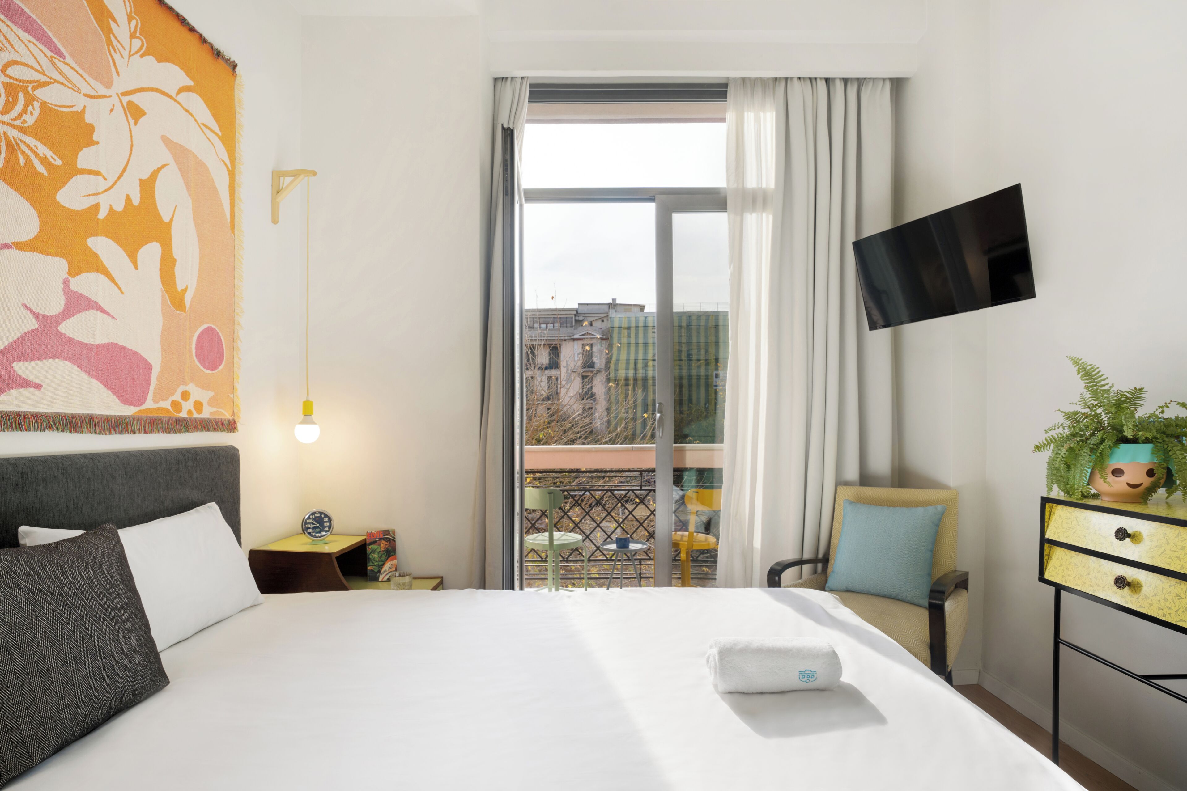 A hotel room with a bed, a TV mounted on the wall, a balcony with a view of buildings, and a plant on a side table.