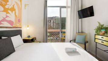 A hotel room with a bed, a TV mounted on the wall, a balcony with a view of buildings, and a plant on a side table.