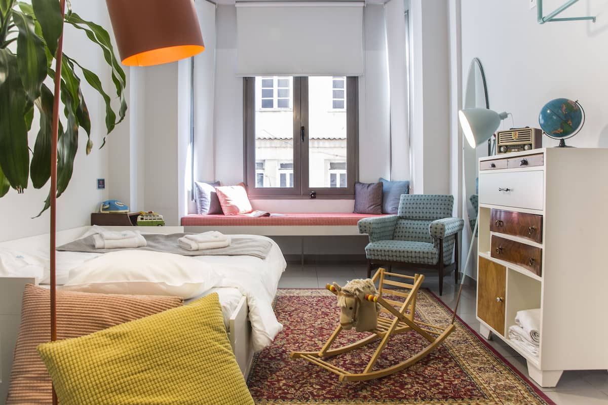 A bedroom with a bed, a rocking chair, a desk, a chair, a wardrobe, a window, and a plant.