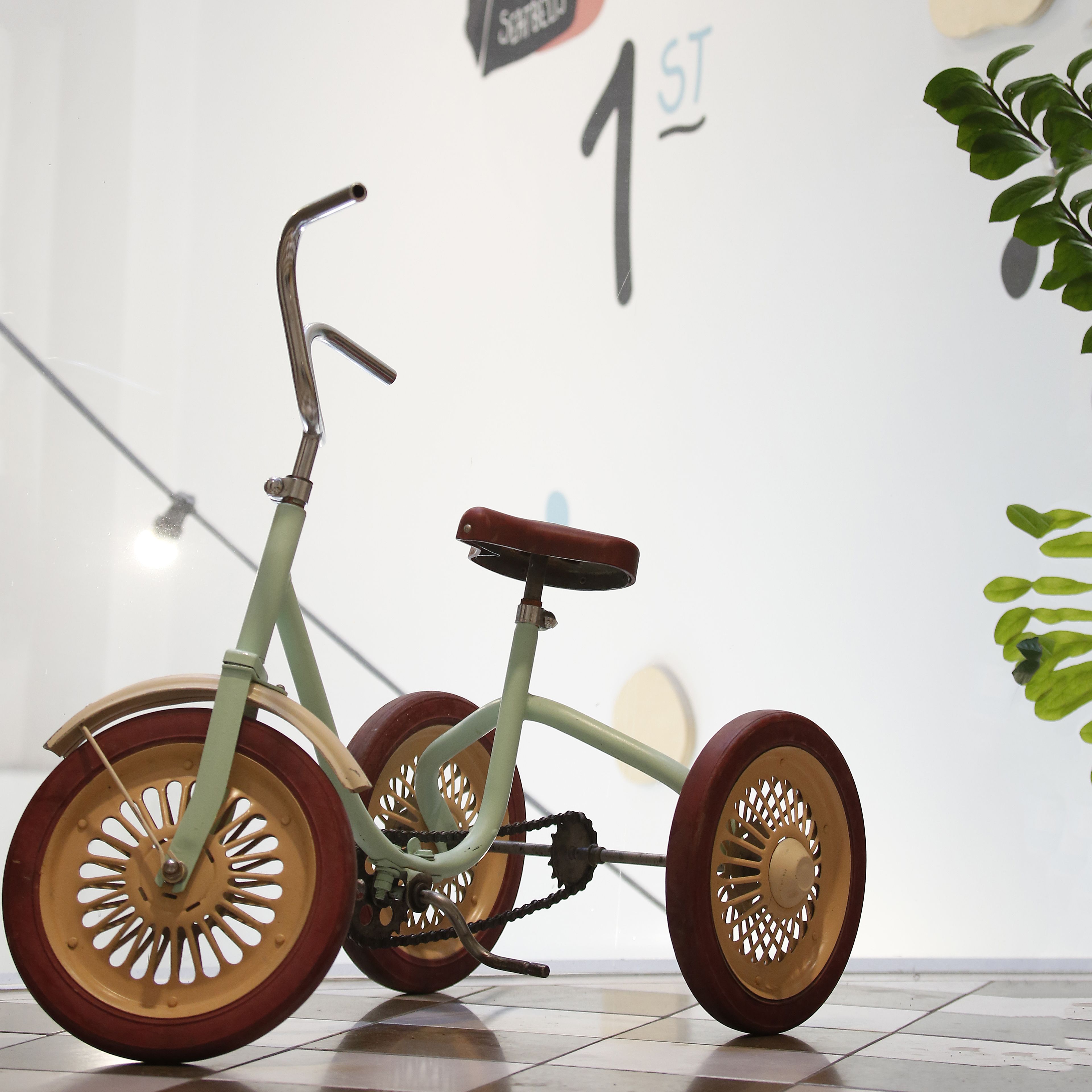 Un triciclo con estructura verde y ruedas de madera, ubicado en un espacio interior con una pared blanca y un cartel al fondo.