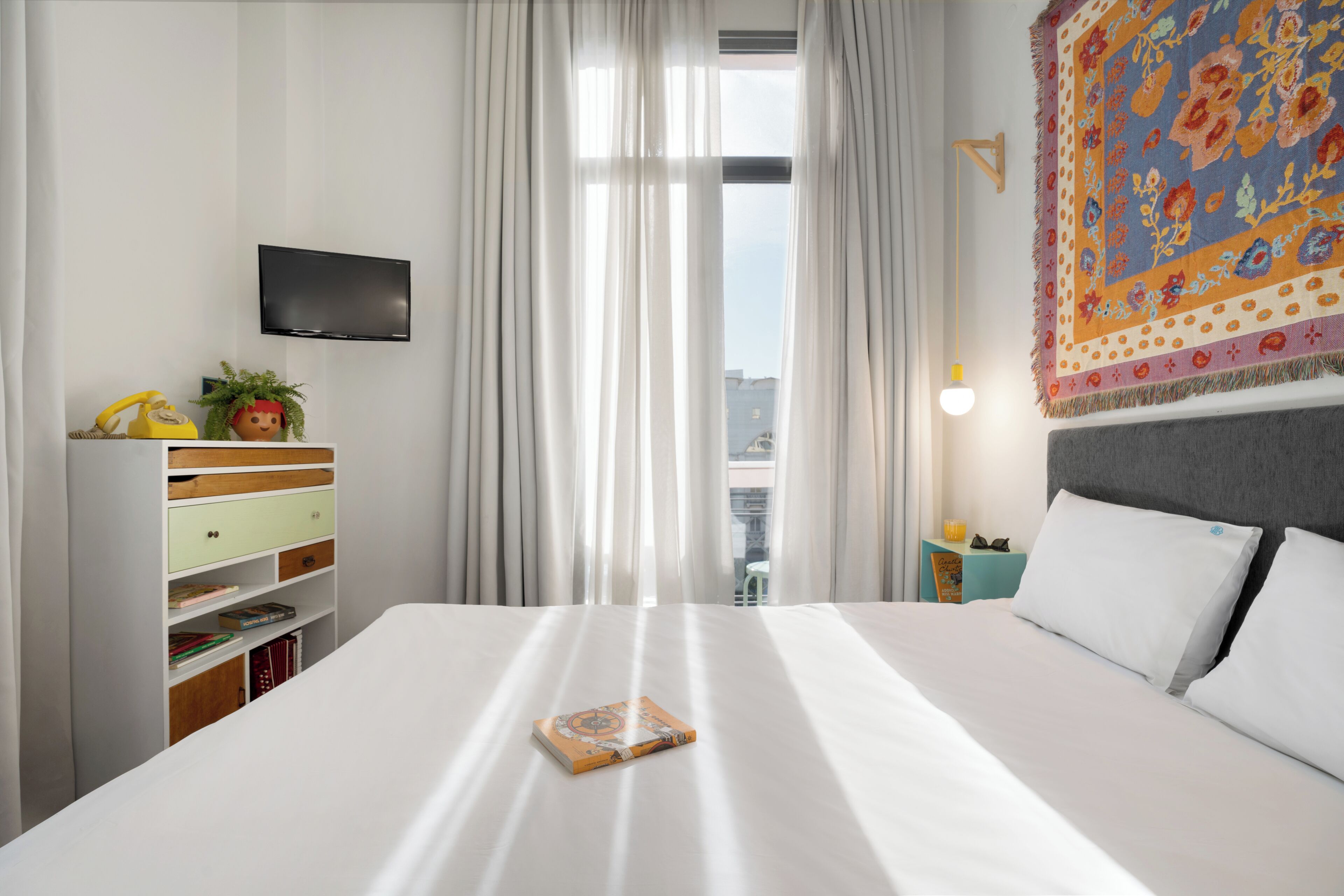 A hotel room with a large bed, a flat-screen TV mounted on the wall, a bookshelf, and a view of the city through sheer curtains.