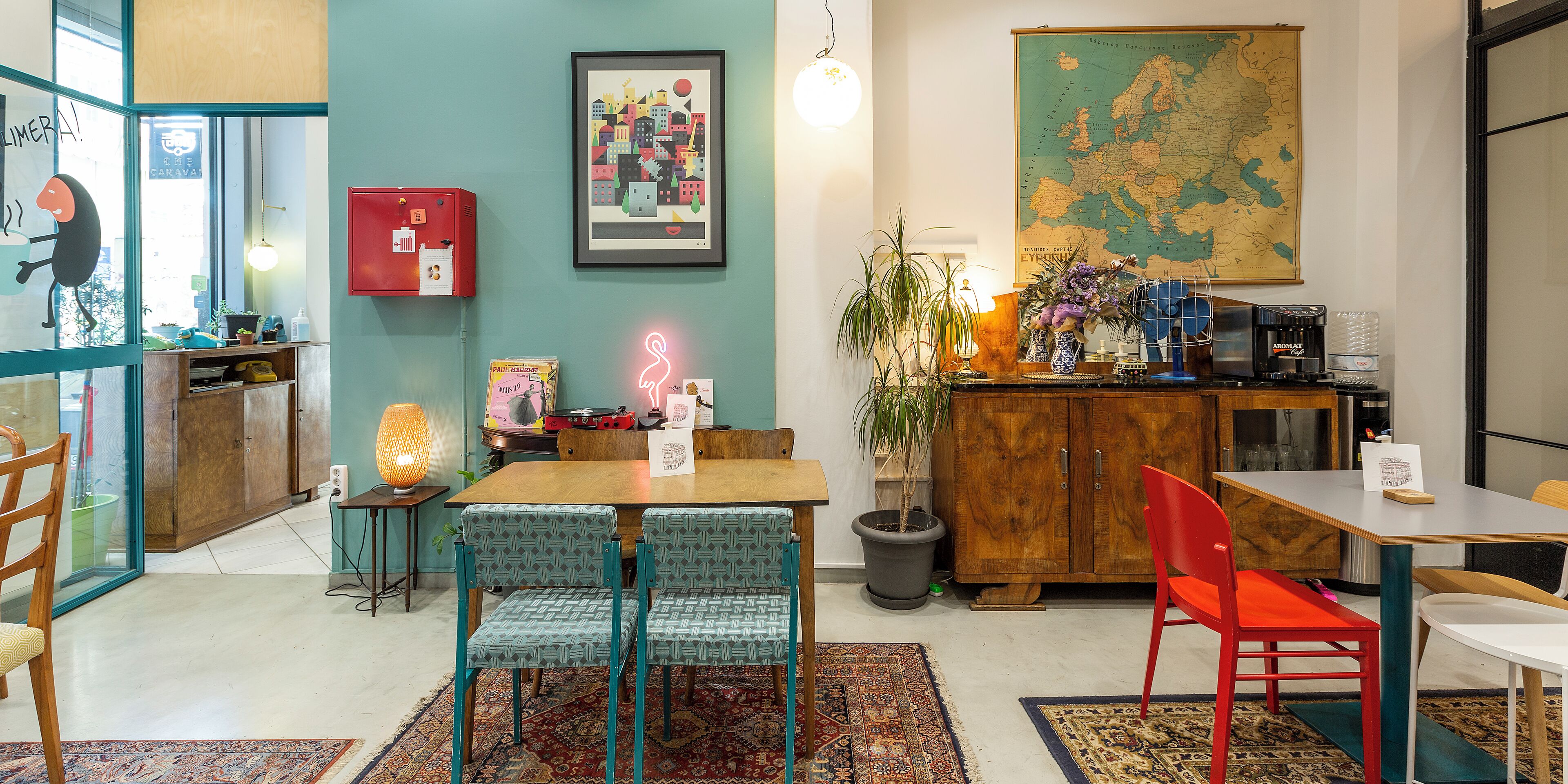 A cozy cafe with a wooden table, two patterned chairs, a red chair, a map on the wall, and a coffee machine.
