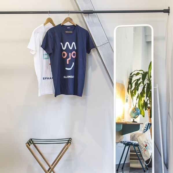 Una habitación con un perchero que sostiene dos camisetas, un espejo, una planta y una silla.