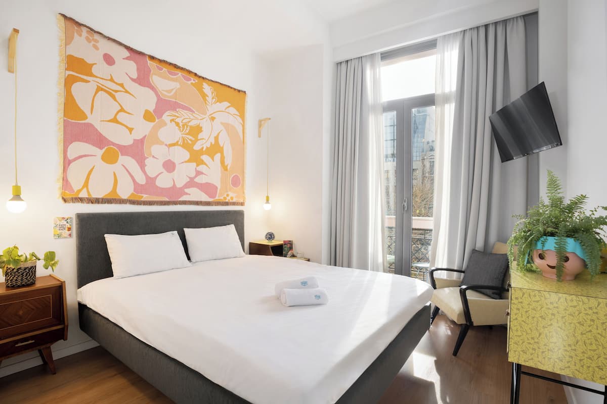 A hotel room with a large bed, a flat-screen TV, a desk with a potted plant, and a window with curtains.
