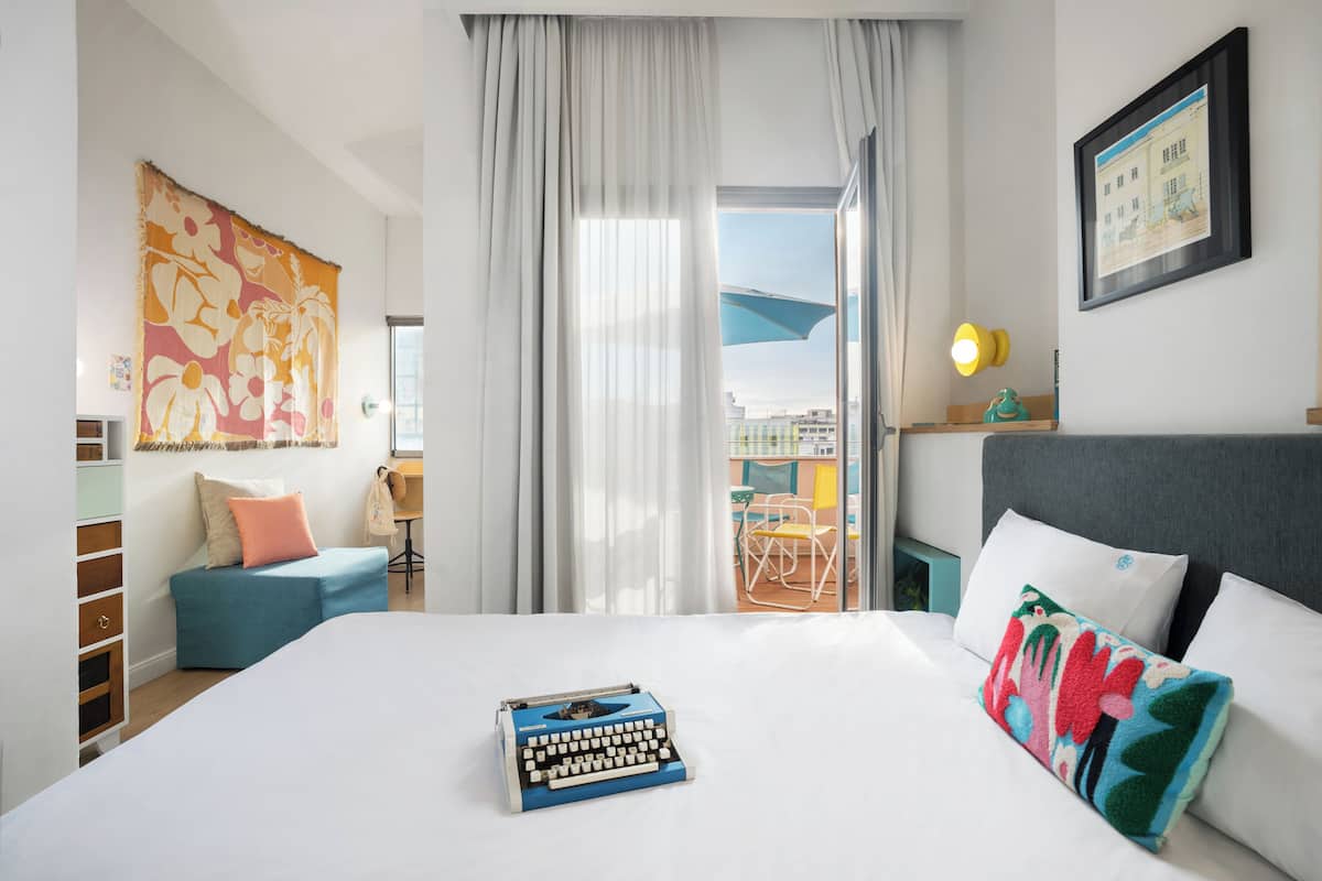 A hotel room with a bed, a desk, a chair, a window with curtains, and a view of the outside.