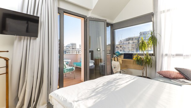 A modern hotel room with a large bed, a TV, a balcony with a view of the city, and a potted plant.