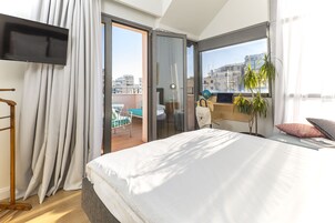 A modern hotel room with a large bed, a TV, a balcony with a view of the city, and a potted plant.