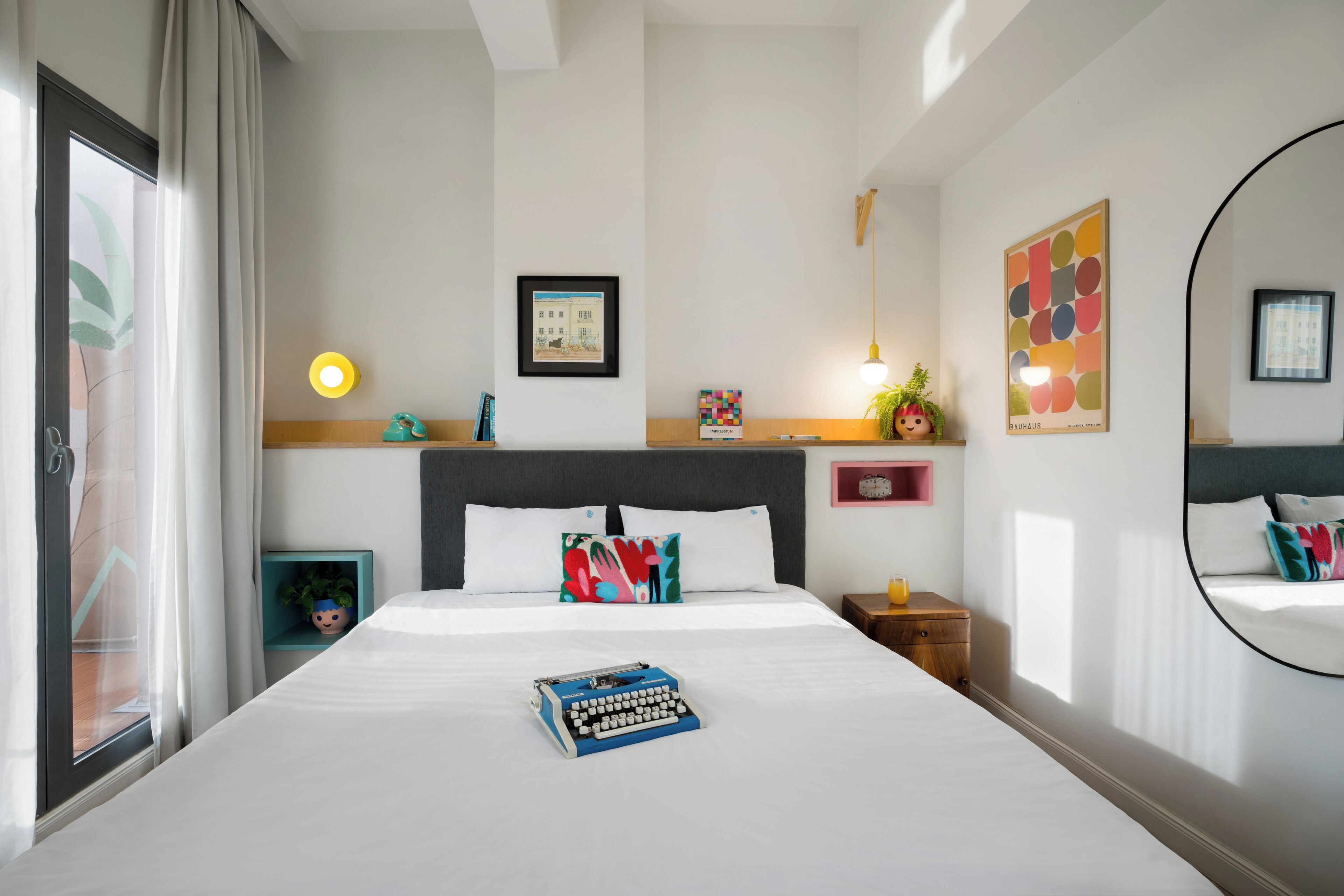 Une chambre d’hôtel moderne avec un grand lit, un bureau, une chaise, une petite table, une plante en pot et une fenêtre avec des rideaux.