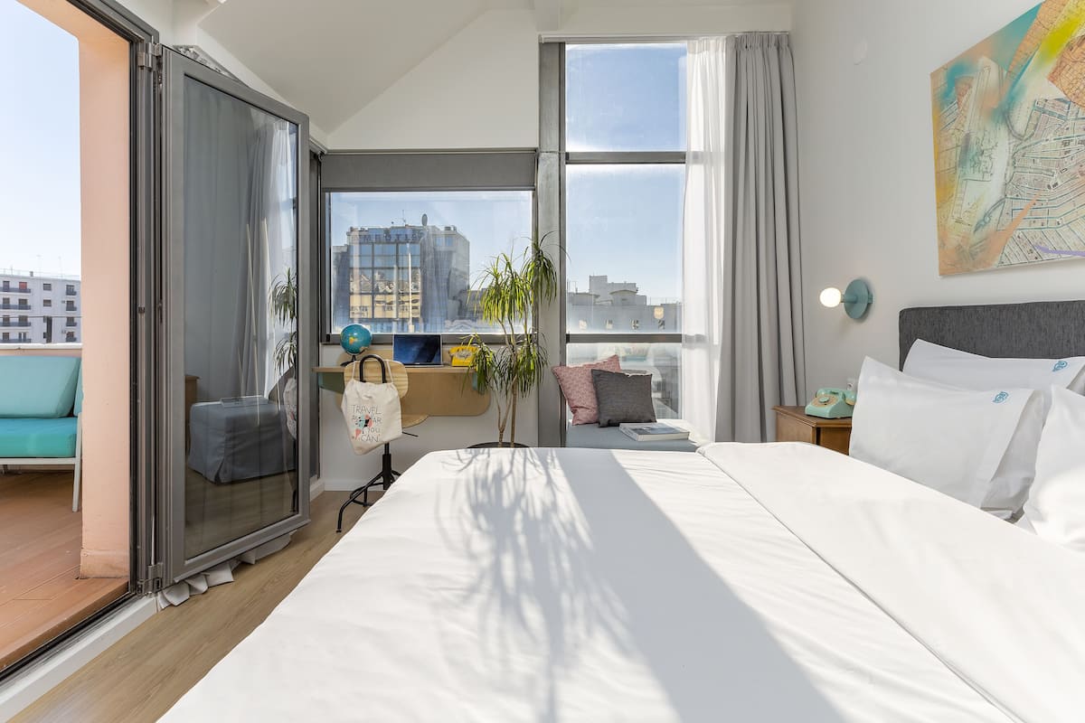 A modern hotel room with a large bed, a desk with a globe, a plant, and a city view through the window.