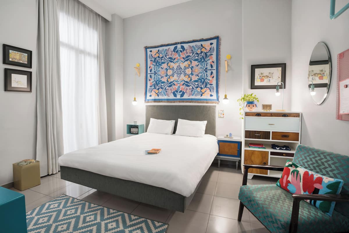 A modern hotel room with a large bed, a desk, a chair, and a decorative tapestry.