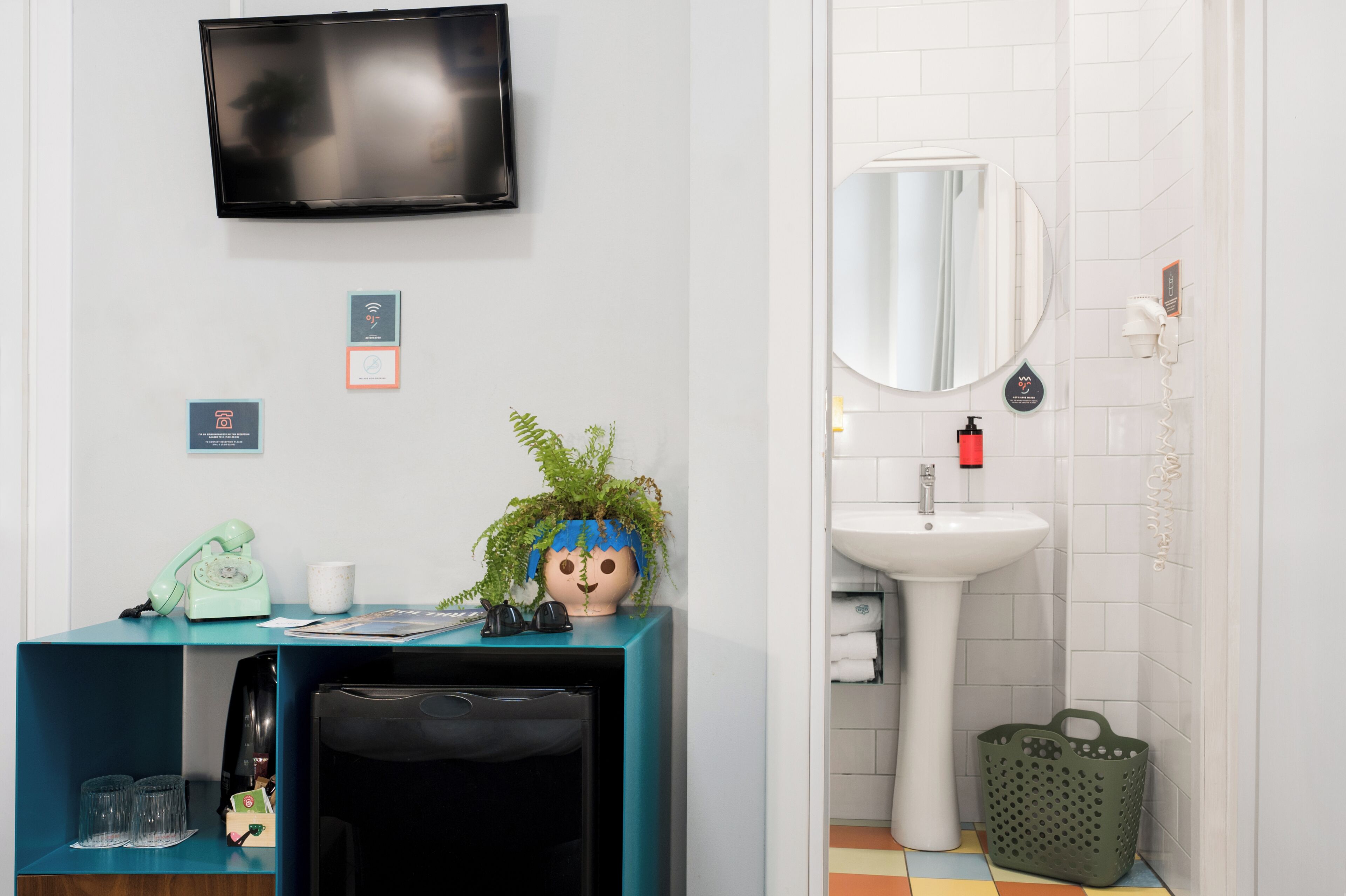 A modern bathroom with a sink, mirror, and toilet. There is a television mounted on the wall, a green basket, and a small plant on the counter.
