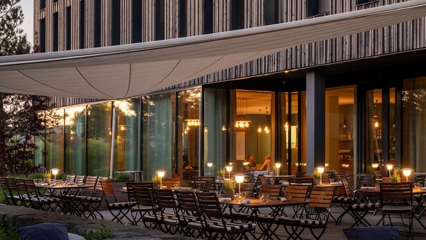 Un área de comedor al aire libre con mesas y sillas, un hogar para fogata y un edificio moderno con líneas verticales y ventanas amplias.