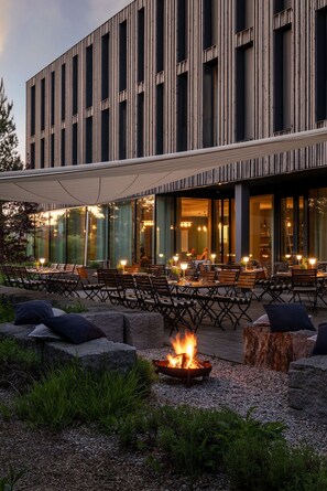 An outdoor dining area with tables and chairs, a fire pit, and a modern building with vertical lines and large windows.