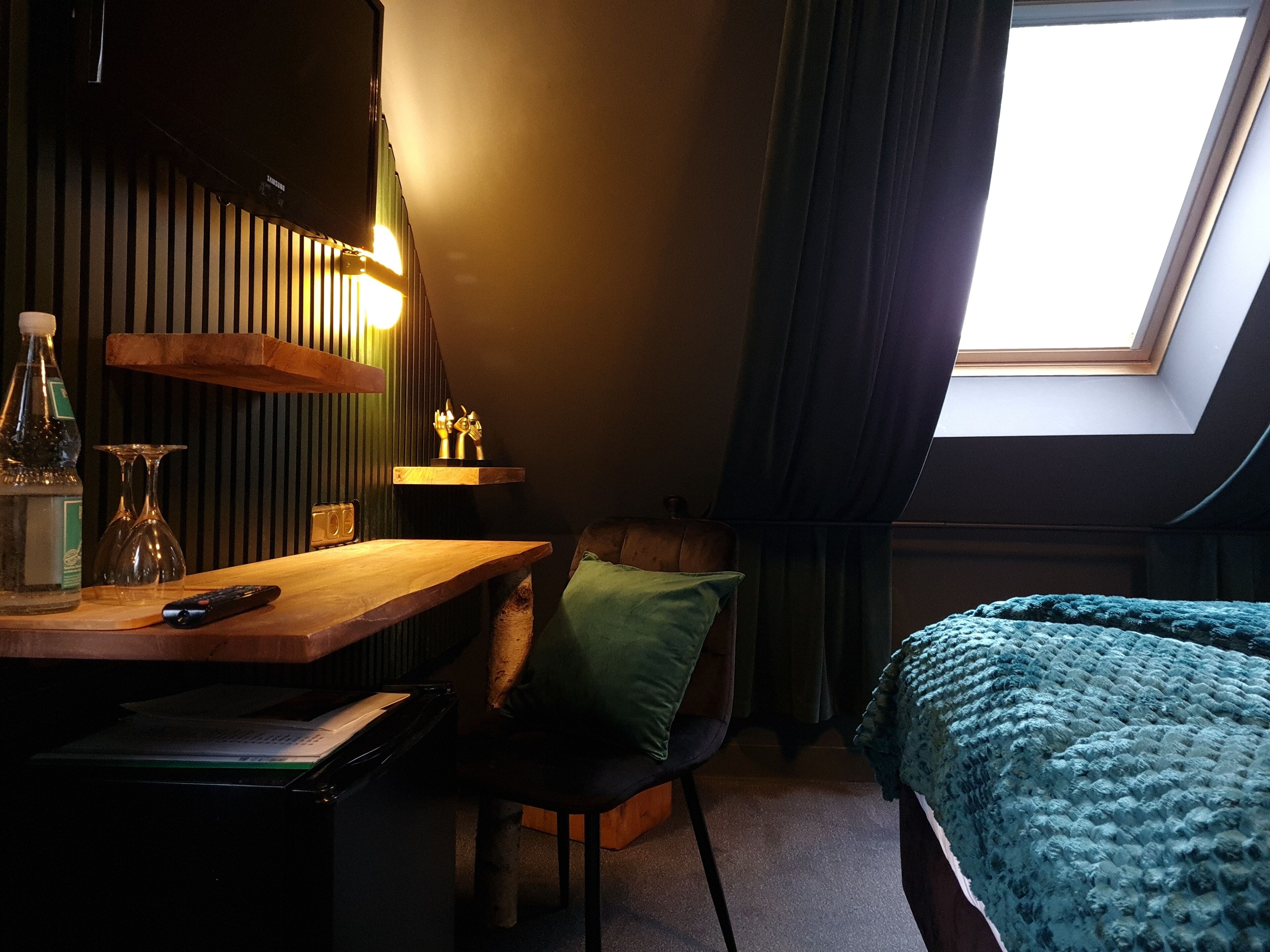 A cozy room with a wooden desk, a chair, a bed with a blue blanket, a skylight, and a green wall.