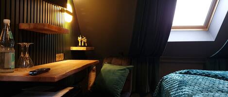 A cozy room with a wooden desk, a chair, a bed with a blue blanket, a skylight, and a green wall.