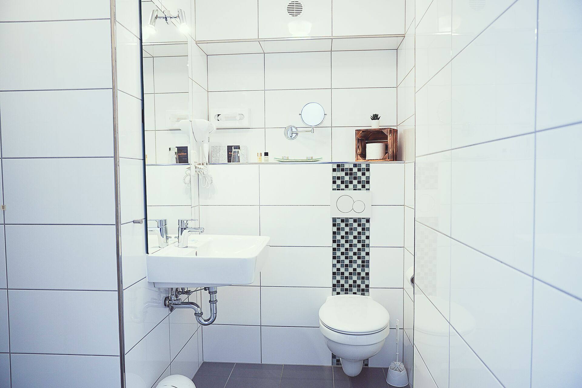 A bathroom with white tiles, a white sink, a toilet, and a small shelf with toiletries.