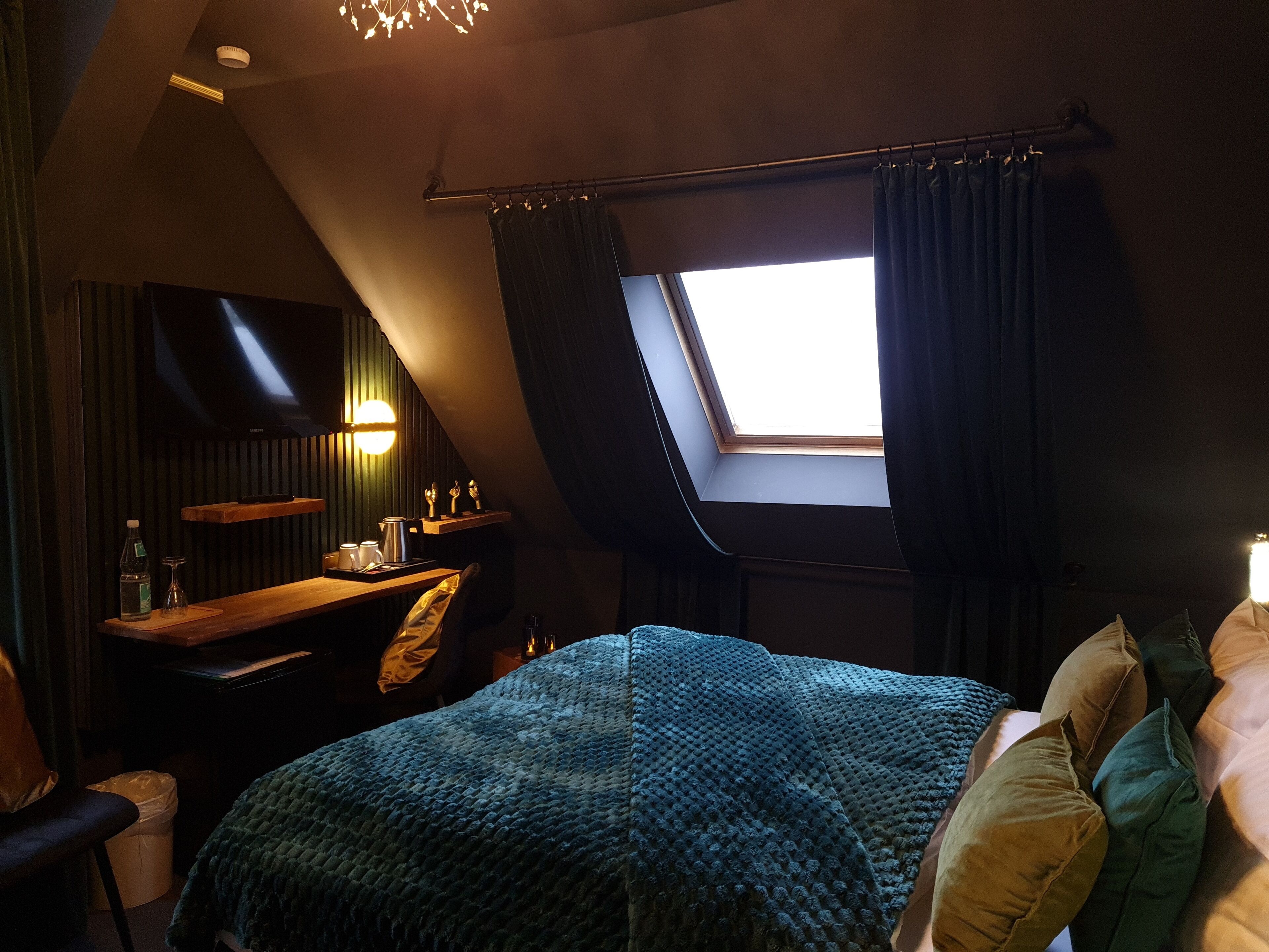 Ein gemütliches Schlafzimmer mit schrägverdeckter Decke, einem Oberlicht, einem Bett mit blauer Decke, einem Holztisch mit Stuhl und einem flachbildfernseher an der Wand.