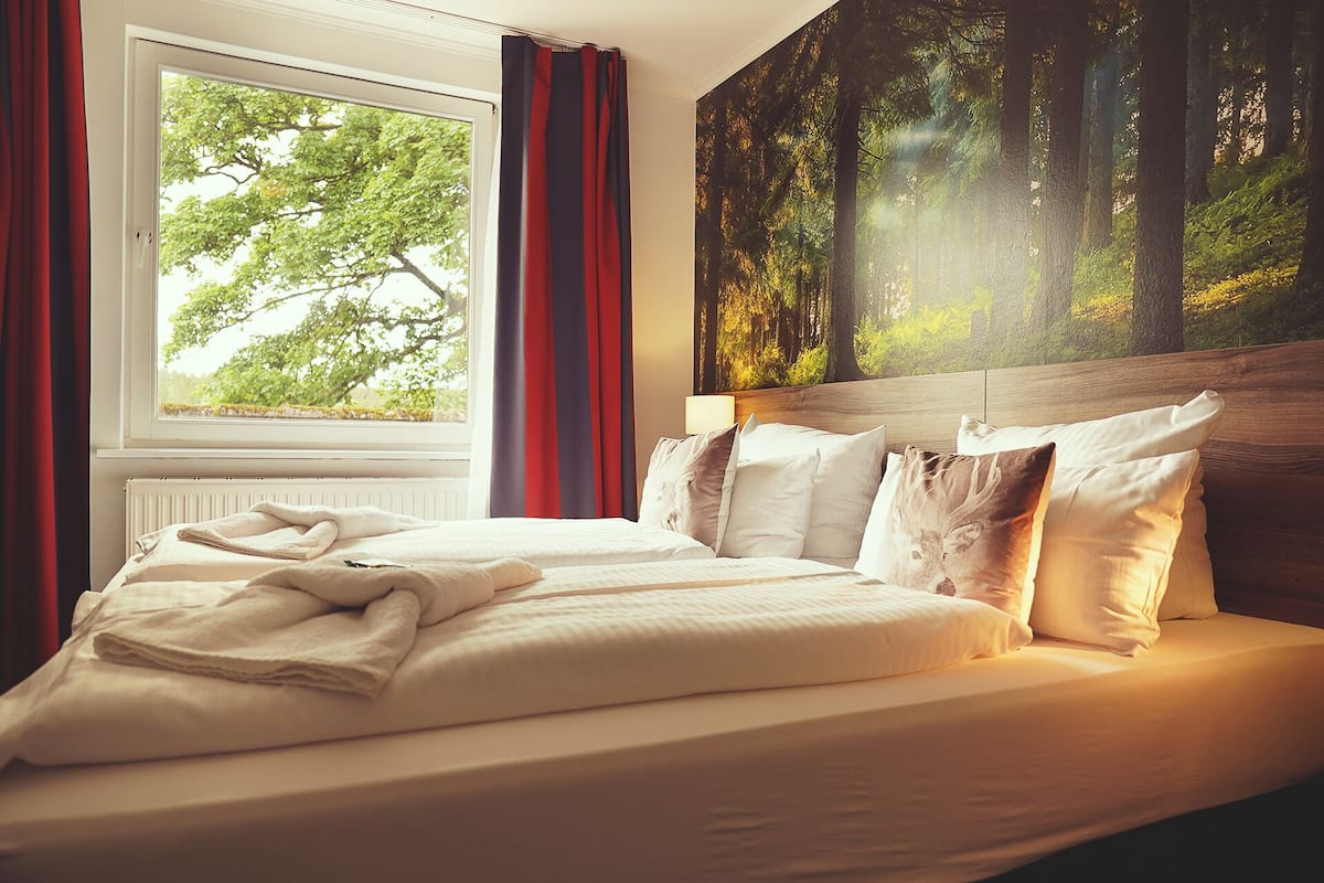 A hotel room with a large bed, a view of trees through the window, and a forest-themed wall mural.