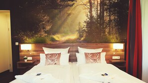 Een tweepersoonsbed met witte lakens en twee kussens, elk met een hertenprint. Twee nachtkastjes met lampen staan ernaast, en aan de muur hangt een groot muurschildering van een boslandschap.
