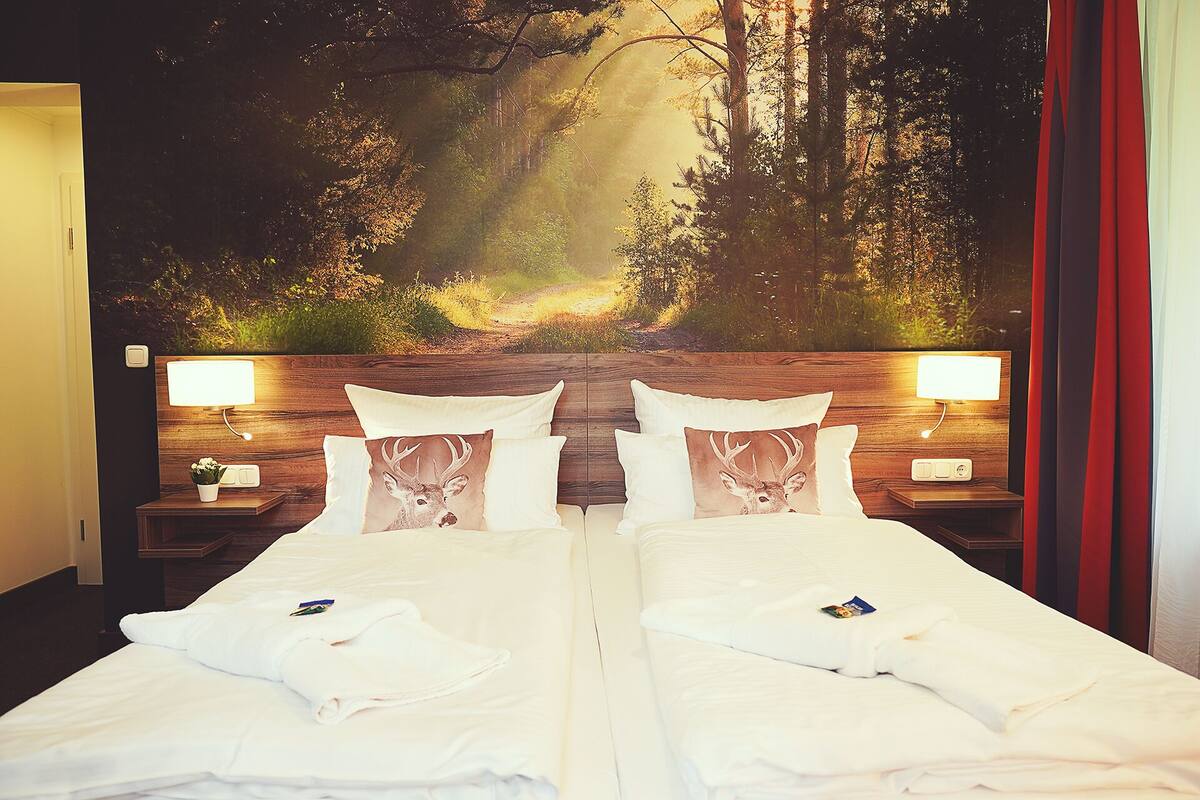 A double bed with white linens and two pillows, each with a deer print. There are two bedside tables with lamps, and a large wall mural of a forest scene.