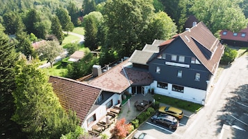 A two-story building with a sign reading "Biga Hotels Hotel Mein Burgpark." It features a parking area with cars, a small outdoor seating area, and is surrounded by trees.