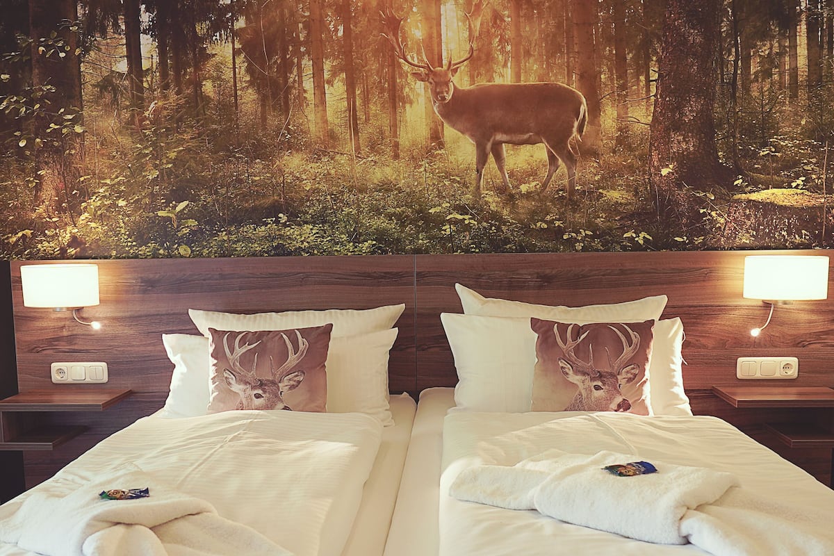A hotel room with two beds, each with a deer-themed pillow, a nightstand with a lamp, and a forest mural on the wall.