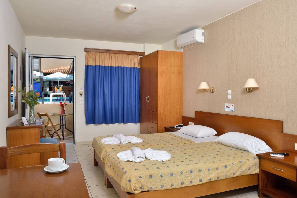 A hotel room with a bed, bedside table, and a desk with a chair. There is a window with curtains, a wall-mounted air conditioner, and a small table with a vase and a cup.