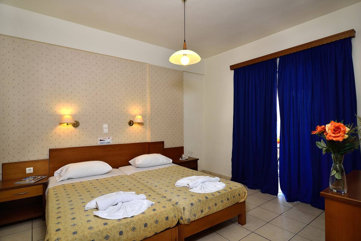 A hotel room with a double bed, two bedside tables, a nightstand, a wall-mounted lamp, a window with blue curtains, and a vase with flowers.