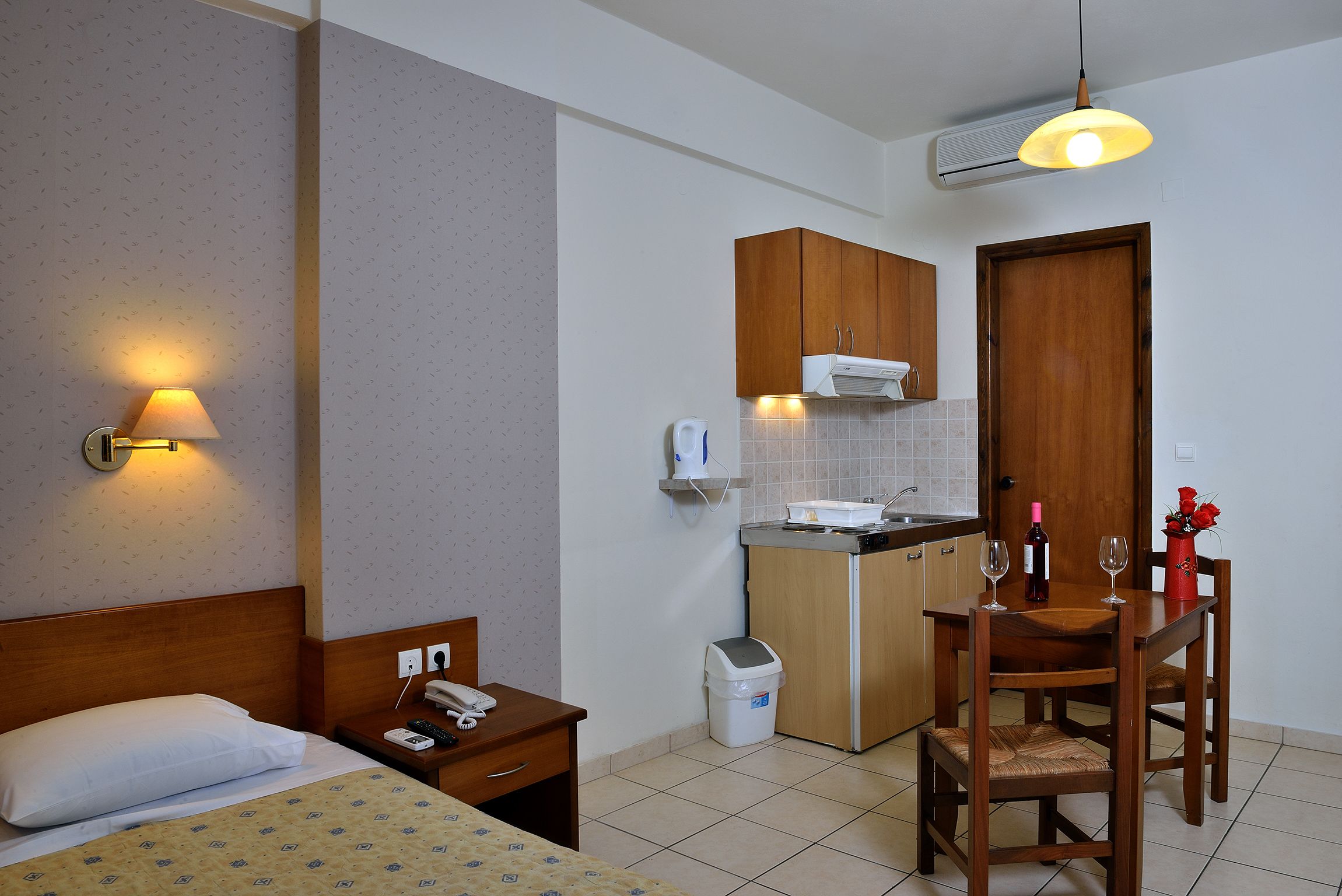 A hotel room with a bed, bedside table, lamp, and a small kitchenette with a table, chairs, and a vase of flowers.