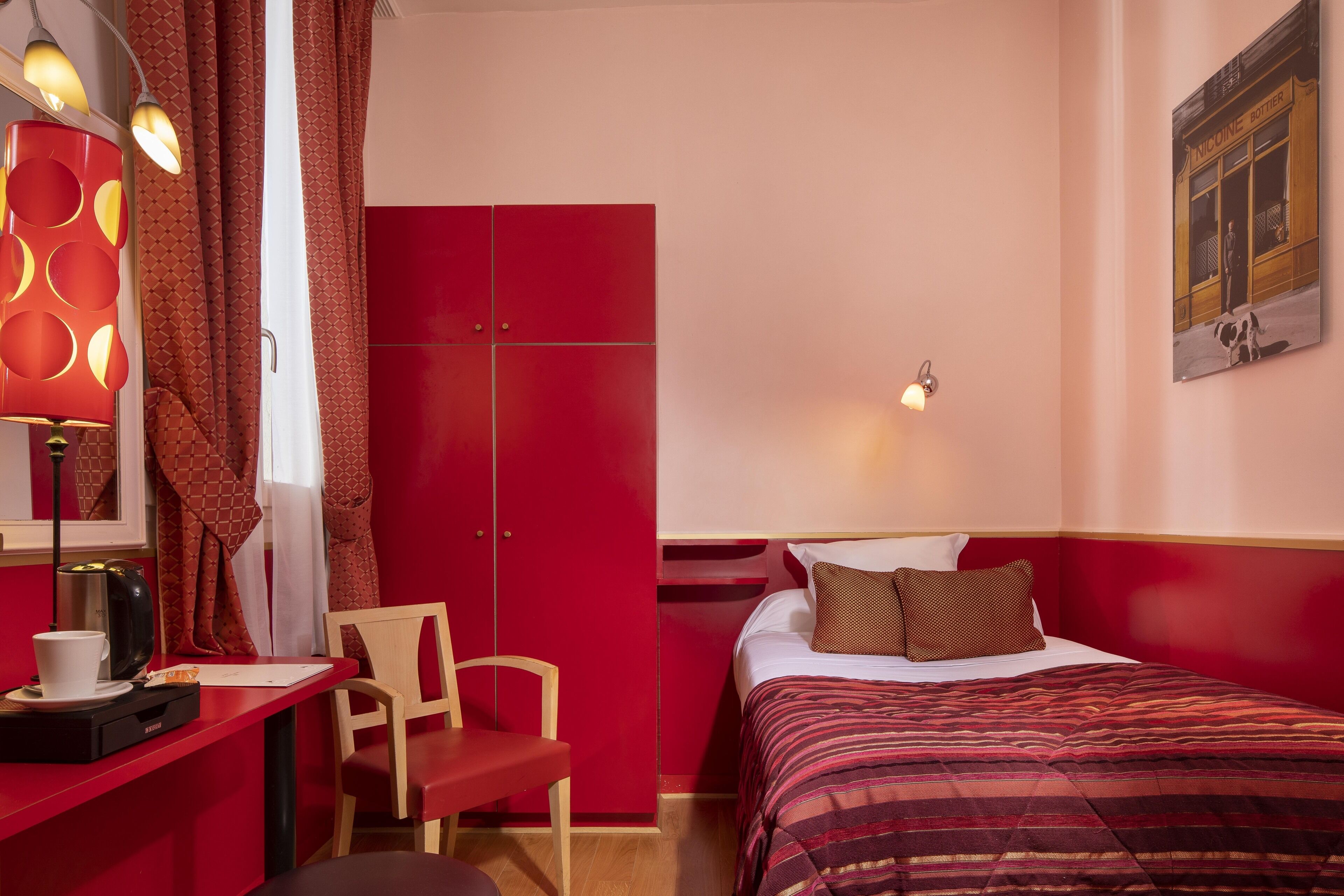 A hotel room with a red wardrobe, a bed with a striped blanket, a desk with a coffee cup, and a wall-mounted picture.