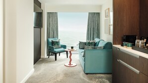 Una sala de estar moderna con vistas al mar, un sofá, dos sillones, una mesita y una zona de cocina con armarios y electrodomésticos.
