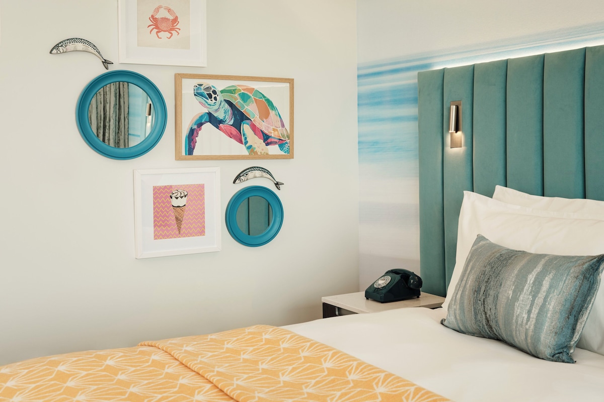 A neatly arranged bedroom with a teal headboard, a bed with white and gray pillows, and wall-mounted artwork including a turtle and a crab.