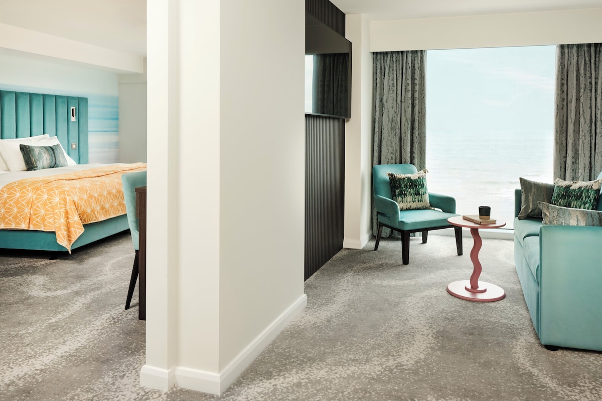 A modern hotel room with a bed, a sofa, a chair, and a small table. There is a large window with curtains and a view of the ocean.