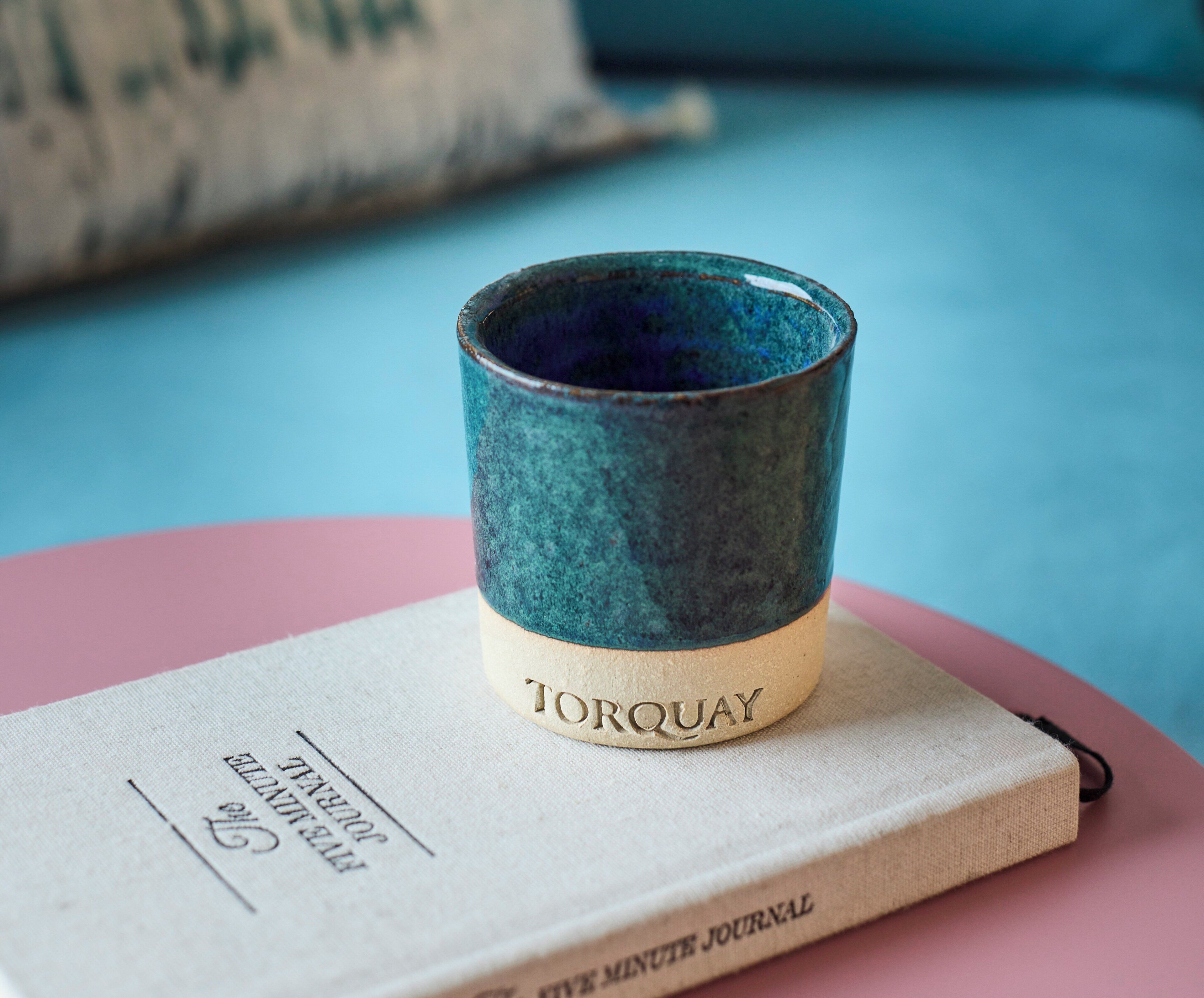 A ceramic mug labeled 'TORQUAY' on a book titled 'MINUTE JOURNAL' on a pink surface.