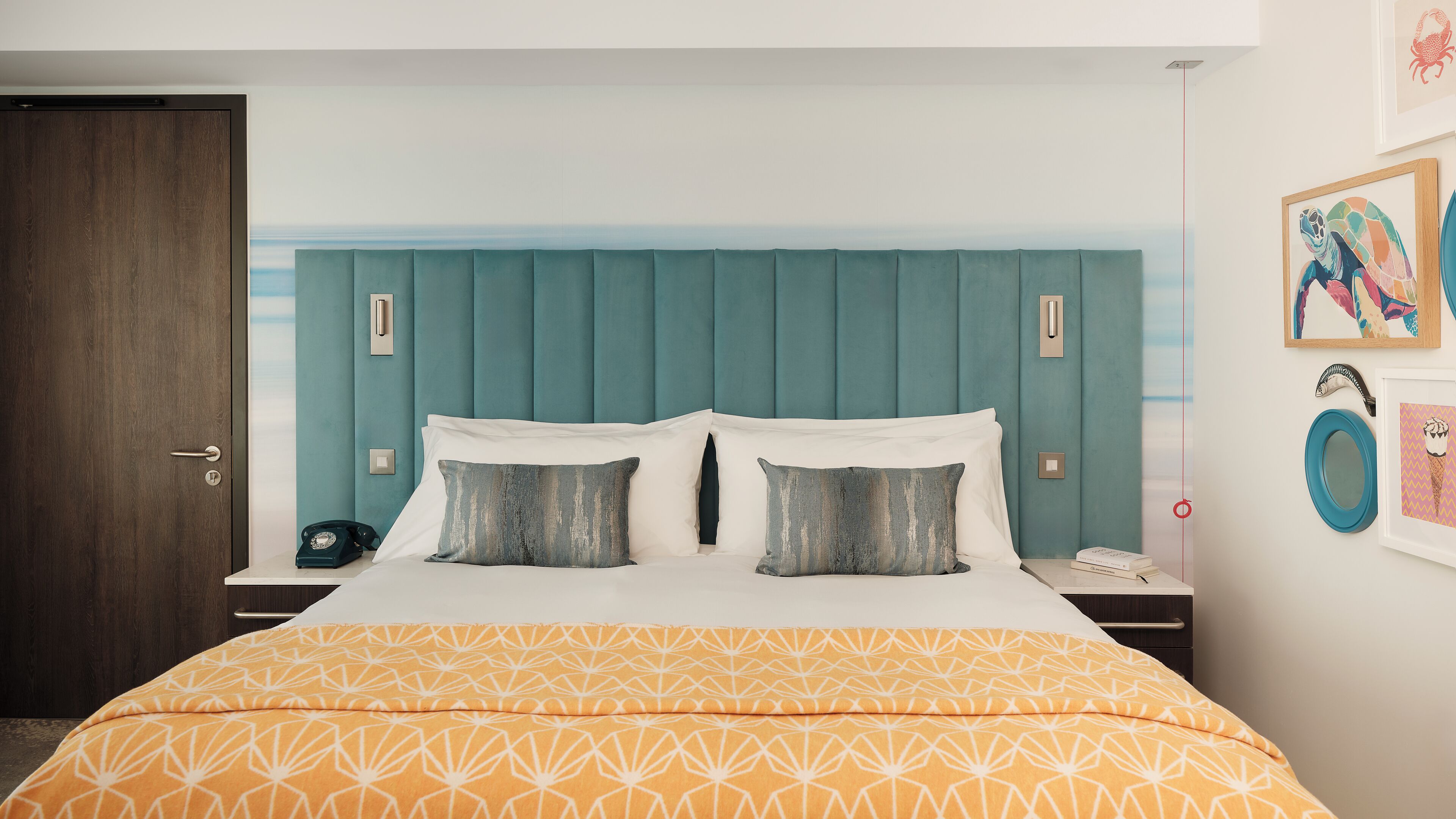 Habitación de hotel con tablero de cabecera color turquesa, colcha con estampado geométrico y mesita de noche con teléfono y un libro.