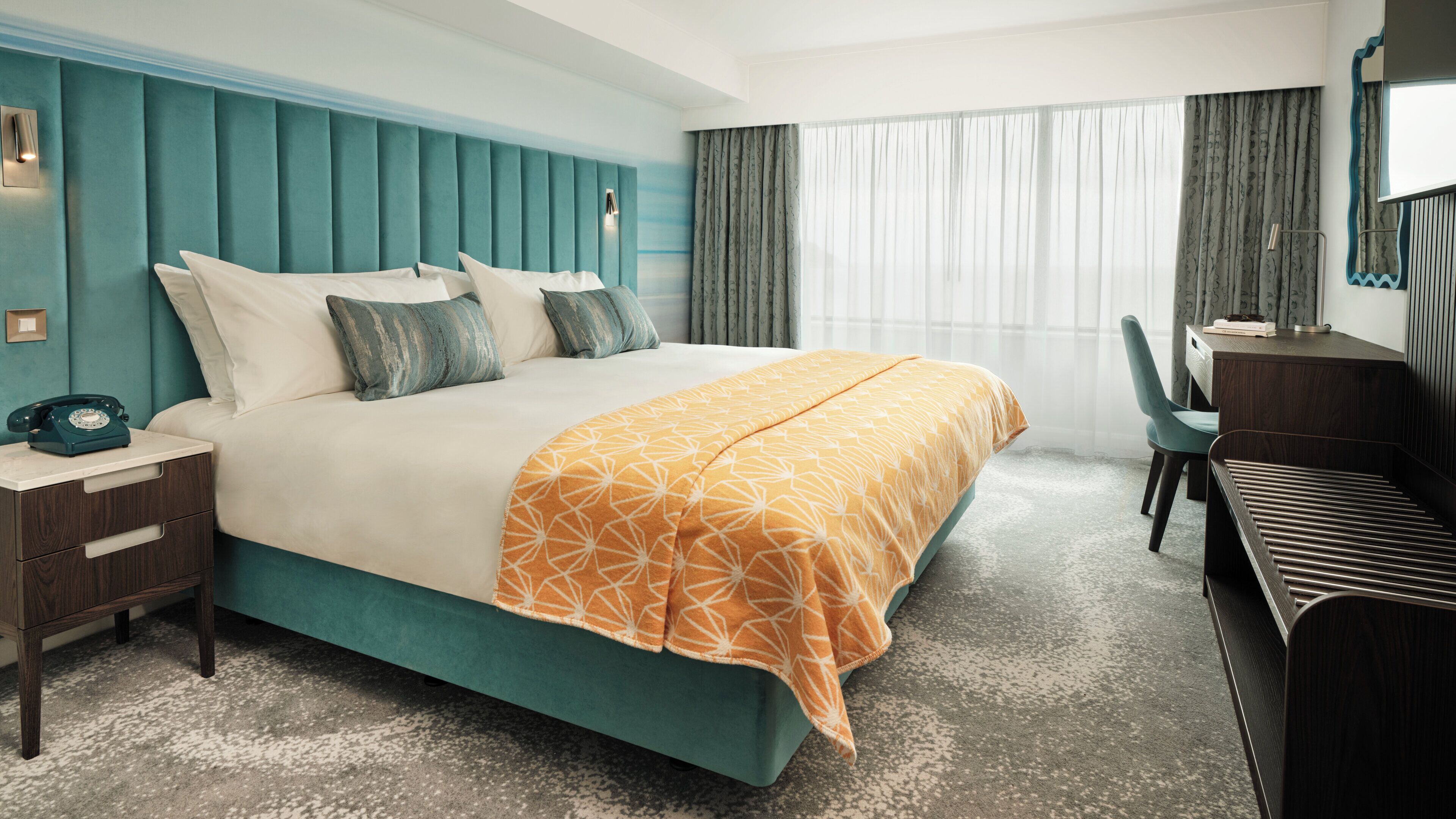 A hotel room with a teal-colored headboard, a bed with white and teal bedding, a desk with a chair, and a large window with sheer curtains.