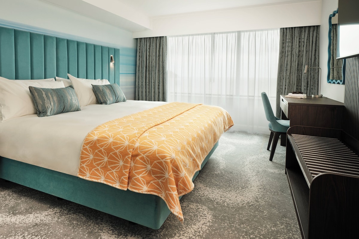 A hotel room with a teal-colored headboard, a bed with white and teal bedding, a desk with a chair, and a large window with sheer curtains.