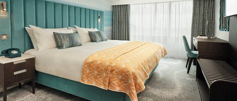 A hotel room with a teal-colored headboard, a bed with white and teal bedding, a desk with a chair, and a large window with sheer curtains.